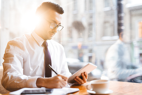 Young Middle Eastern business executive working at window table in cafe by rays of warm sunset light: holding tablet while making notes with pen on paper