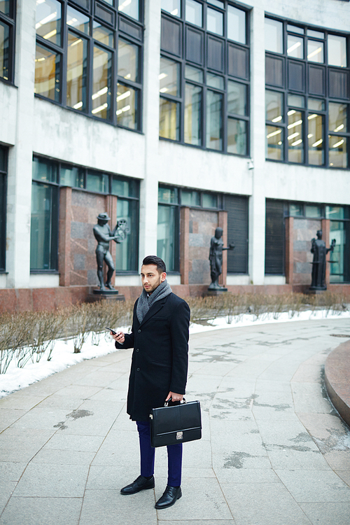 Portrait of stylish Middle-Eastern businessman holding smartphone and briefcase against background of modern building in city