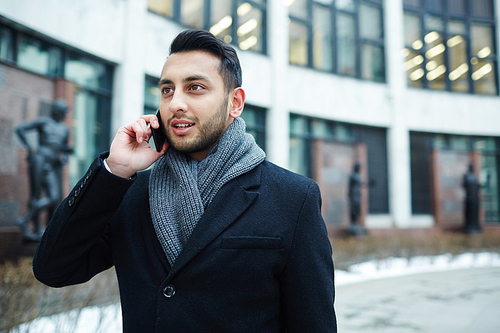 Portrait of stylish Middle-Eastern businessman speaking by phone against background of modern building in city