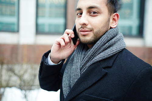 Portrait of young Middle-Eastern businessman speaking by phone against background of modern building in city