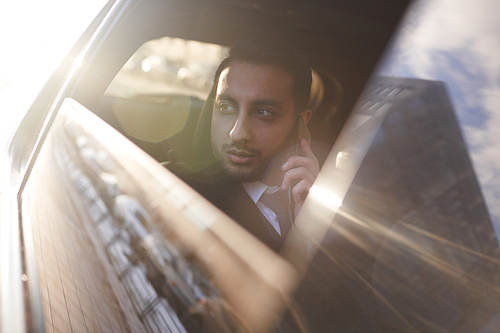 Portrait of handsome Middle-Eastern businessman riding in backseat of car speaking by phone, shot out of window