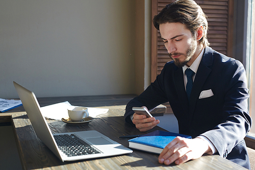 Portrait of handsome elegant businessman working with smartphone and laptop at wooden sunlit table