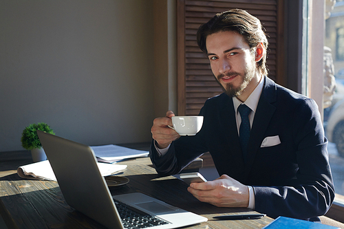 Portrait of young handsome businessman working with smartphone and laptop at wooden sunlit table during coffee break,  and smiling charmingly