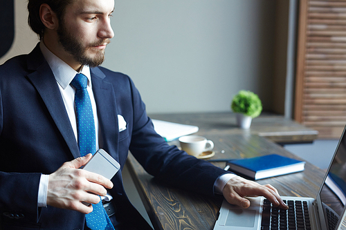 Portrait of handsome elegant businessman busy working with laptop and smartphone at wooden table in cafe