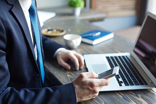 Side view closeup of elegant modern businessman during coffee break: holding  smartphone and typing text messages while working with laptop in cafe