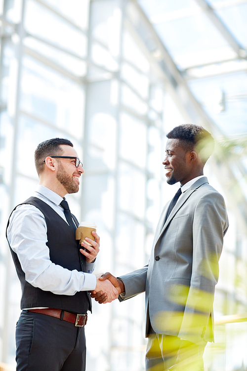 Portrait of smiling businessman greeting African-American partner by handshake standing in modern glass hall of office building