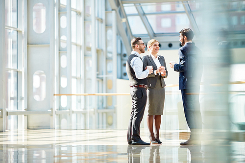 Group of modern business people chatting during coffee break  standing in sunlit glass hall of office building