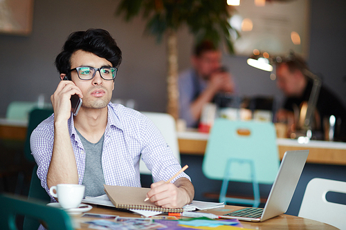 Portrait of young casual creative man calling by phone while sitting at table in cafe making notes