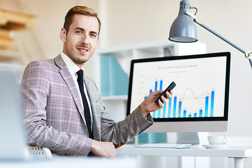 Successful young banker with smartphone sitting by his workplace