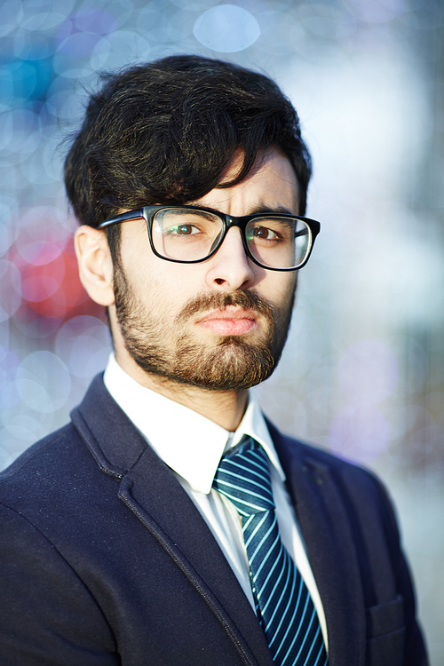 Head and shoulders portrait of young confident Middle-Eastern businessman wearing glasses and elegant suit   with serious expression against blurred office background