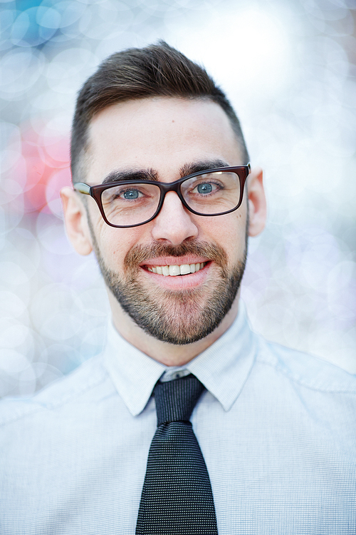 Head and shoulders portrait of young confident bearded businessman wearing glasses and white shirt with tie smiling cheerfully against blurred office background