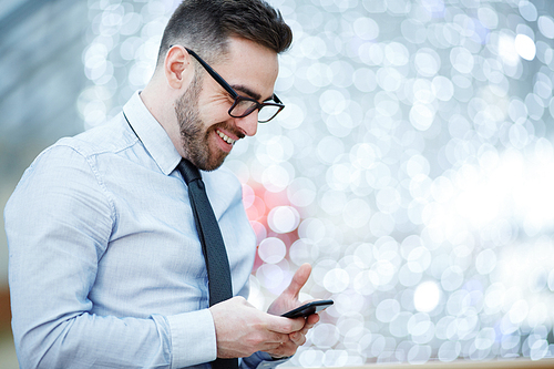 Side view portrait of modern businessman wearing glasses, shirt and tie smiling joyfully while reading text message from smartphone against blurred office background