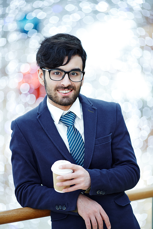 Portrait of handsome Middle Eastern man wearing glasses and elegant formal suit smiling confidently at camera while standing leaning on railing in office building holding coffee cup against blurred background