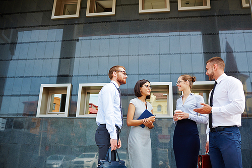 Group of young successful business colleagues chatting and smiling outside modern office building