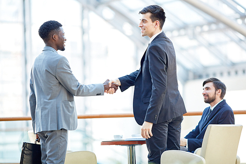 Portrait of smiling successful businessman greeting African-American man in meeting shaking hands at table in office building