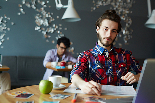 Young creative bearded businessman wearing long hair and colorful casual shirt looking confidently at camera while working with laptop at table in cafe, other people in background