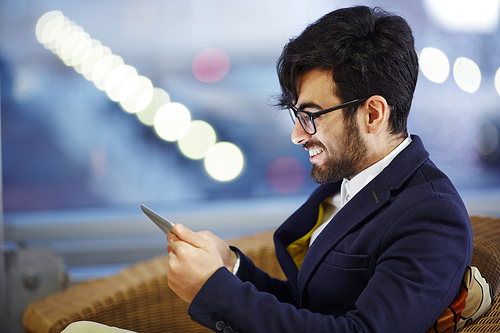 Side view portrait of young handsome middle eastern businessman sitting in leather armchair using smartphone and smiling reading text message