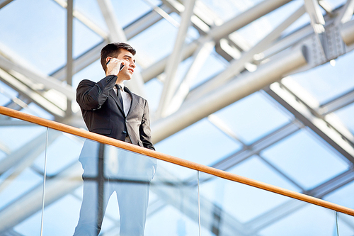 Low angle portrait of powerful young businessman talking by smartphone standing on glass balcony of modern office building