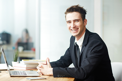 Happy manager in suit sitting by workplace in front of laptop in office