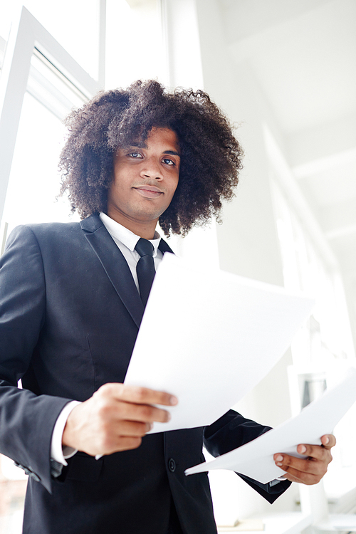 Confident financier with papers standing by office window