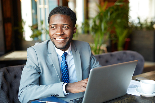 Portrait of handsome African- American businessman wearing formal suit smiling cheerfully while working with laptop in cafe