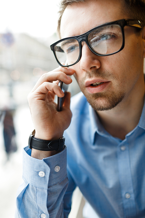 Portrait of young businessman wearing big black glasses discussing work speaking by mobile phone at window in cafe