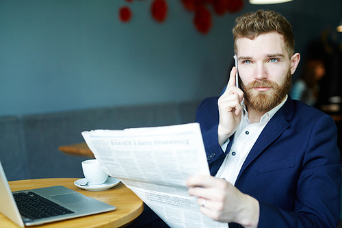 Portrait of handsome young businessman speaking by smartphone while reading newspaper  in cafe