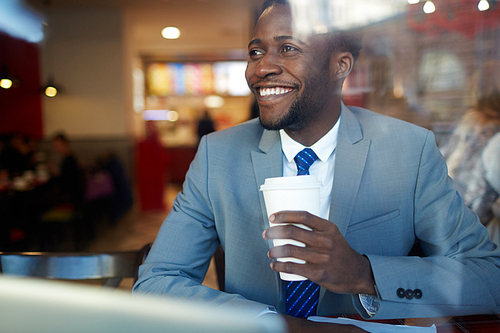 Portrait of cheerful African ?American businessman drinking coffee from disposable cup and looking away smiling  in coffee shop, shot behind glass window