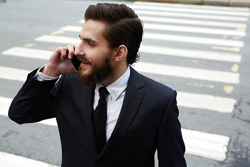 Young busy man with smartphone speaking while crossing street