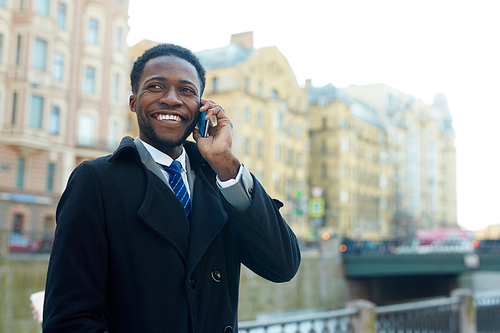 Portrait of two businessman in autumn street of city, African ?American man smiling cheerfully while talking by phone standing on riverbank and wearing coat