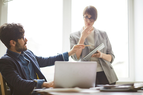 Young successful middle eastern man looking up from laptop to  consult young colleague standing by him with tablet, pointing at it