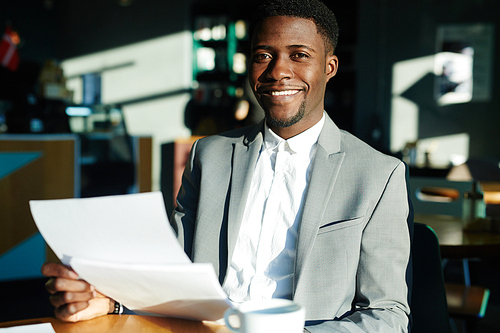 Portrait of successful African-American businessman sitting in bright sunlight at table in cafe and working with documents and smiling 