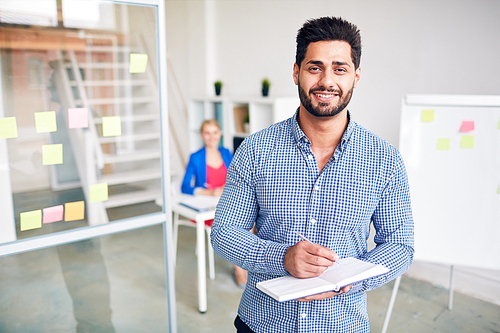 Young smiling manager making working notes in notebook with his colleague on background