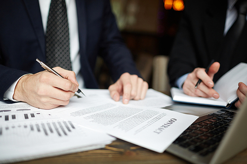 Closeup portrait of two unrecognizable business people wearing black formal suits signing contract papers at table during meeting