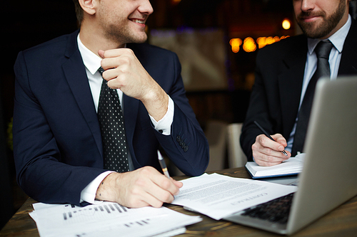 Closeup portrait of two unrecognizable successful business partners wearing black formal suits reviewing income statistics and signing contract papers at table during meeting