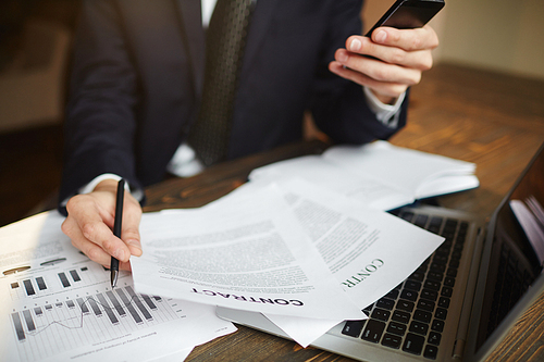 Portrait of busy modern businessman working in office with documentation, contracts and income statistics while using smartphone