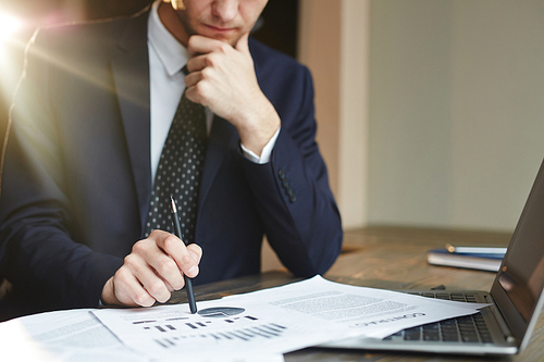 Closeup portrait of unrecognizable successful businessman wearing black formal suit analyzing  finance statistics and contract documents at desk, thinking