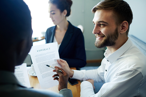 Portrait of young smiling businessman sitting at meeting table with three partners and discussing contract with them