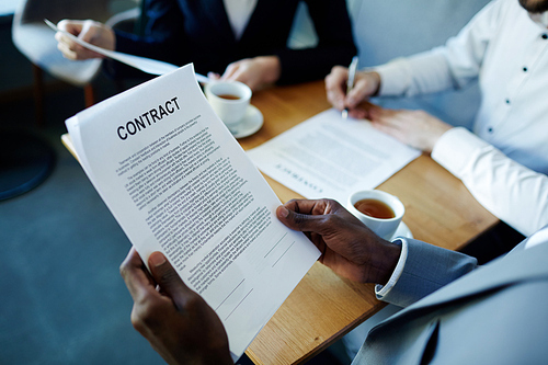 Closeup shot of business contract in hands of African man reading it while sitting with partners at meeting table