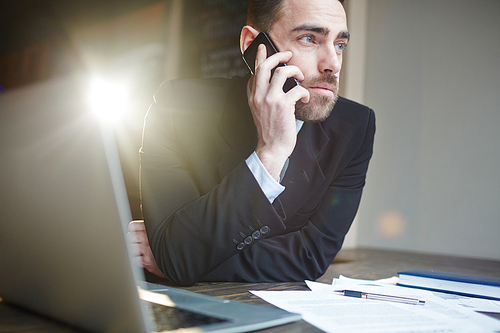 Portrait of modern bearded businessman working in office using laptop and talking by mobile phone against black background with lens flare