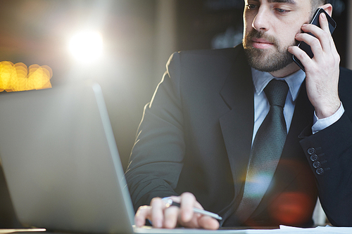 Portrait of modern bearded businessman wearing black formal suit busy working in office using laptop and talking by mobile phone against black background with lens flare