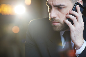 Portrait of modern bearded businessman wearing black formal suit looking serious and busy talking by mobile phone against black background with lens flare