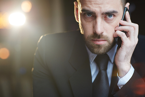 Portrait of modern businessman wearing black formal suit  while busy talking by mobile phone against black background with lens flare