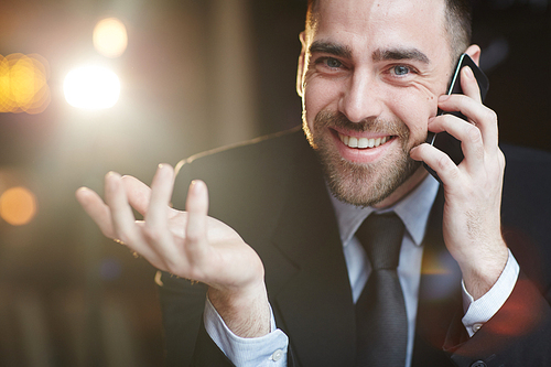 Portrait of successful bearded businessman wearing  formal suit smiling cheerfully to camera and gesturing while talking by mobile phone with  client against black background