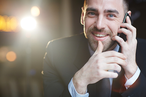 Portrait of modern handsome businessman using smartphone making call and smiling at camera while talking against black background  with lens flare