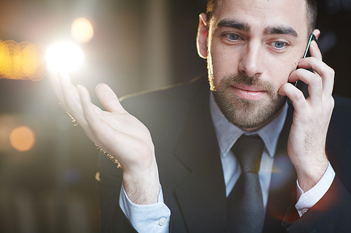 Portrait of successful bearded businessman smiling and gesturing while talking by mobile phone with client against black background  with lens flare