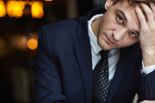 Portrait of young tired businessman wearing formal suit  with serious neutral expression resting head on hand