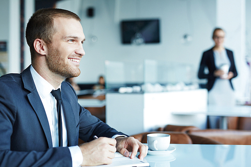 Young handsome businessman  wearing suit smiling while relaxing with cup of coffee during break in restaurant of modern office center, taking time to make notes in planner