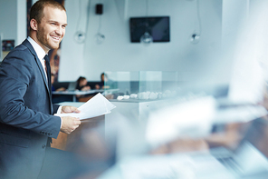 Handsome young businessman wearing formal attire standing up laughing, holding documents during coffee break in modern restaurant hall of office building