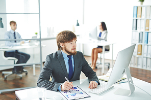 Portrait of young smiling businessman looking at computer screen, checking statistics report while sitting at workplace in modern office with other people blurred in background